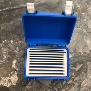 May include: A blue plastic storage box, open to reveal a white interior with dividers. The box has a hinged lid and white latches. The dividers create slots for organising small items. The box is on a marble surface. The box is approximately 15 cm long.