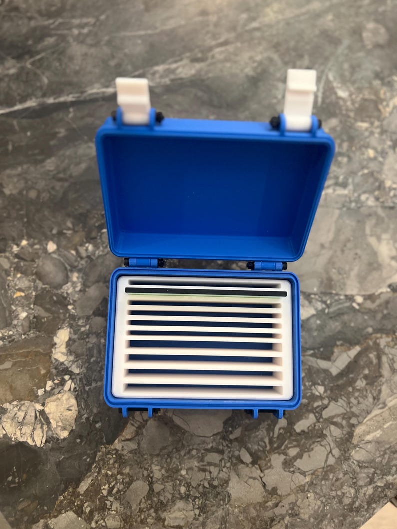 May include: A blue plastic storage box, open to reveal a white interior with dividers. The box has a hinged lid and white latches. The dividers create slots for organising small items. The box is on a marble surface. The box is approximately 15 cm long.