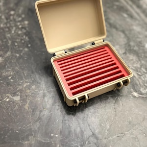 May include: An open, tan-colored, small carrying case with red interior foam and red latches. The case is designed to protect items and has a rectangular shape. The case is on a gray surface.