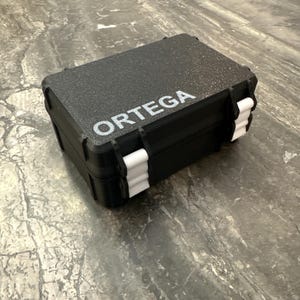 Pu&ograve; includere: Una valigetta rettangolare nera con la scritta "ORTEGA" stampata in bianco. La valigetta ha chiusure bianche e una superficie strutturata. La valigetta &egrave; chiusa e sembra essere realizzata in un materiale resistente.