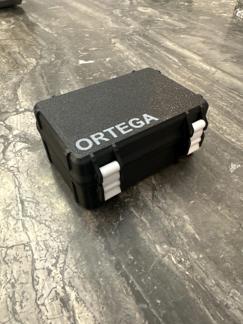 Pu&ograve; includere: Una valigetta rettangolare nera con la scritta "ORTEGA" stampata in bianco. La valigetta ha chiusure bianche e una superficie strutturata. La valigetta &egrave; chiusa e sembra essere realizzata in un materiale resistente.
