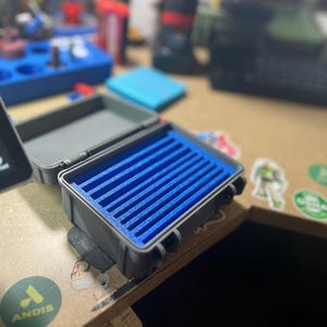 May include: A small, open gray case with a blue foam insert. The case is rectangular and has a hinged lid. The blue foam has multiple grooves. The case is sitting on a wooden surface with stickers.