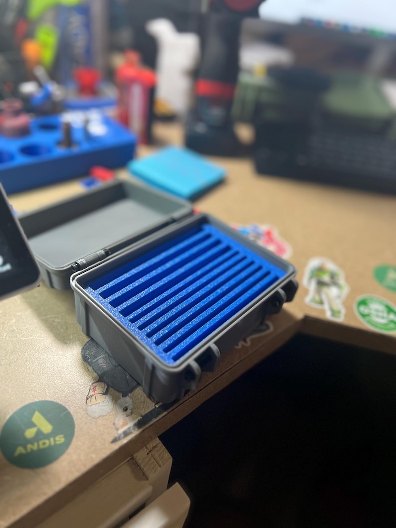 May include: A small, open gray case with a blue foam insert. The case is rectangular and has a hinged lid. The blue foam has multiple grooves. The case is sitting on a wooden surface with stickers.