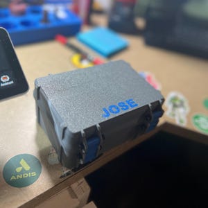 Puede incluir: Una caja gris texturizada con cierres azules y el nombre "JOSE" en letras azules. La caja está cerrada y parece estar hecha de plástico duradero. El fondo está borroso, mostrando otros objetos sobre un escritorio.