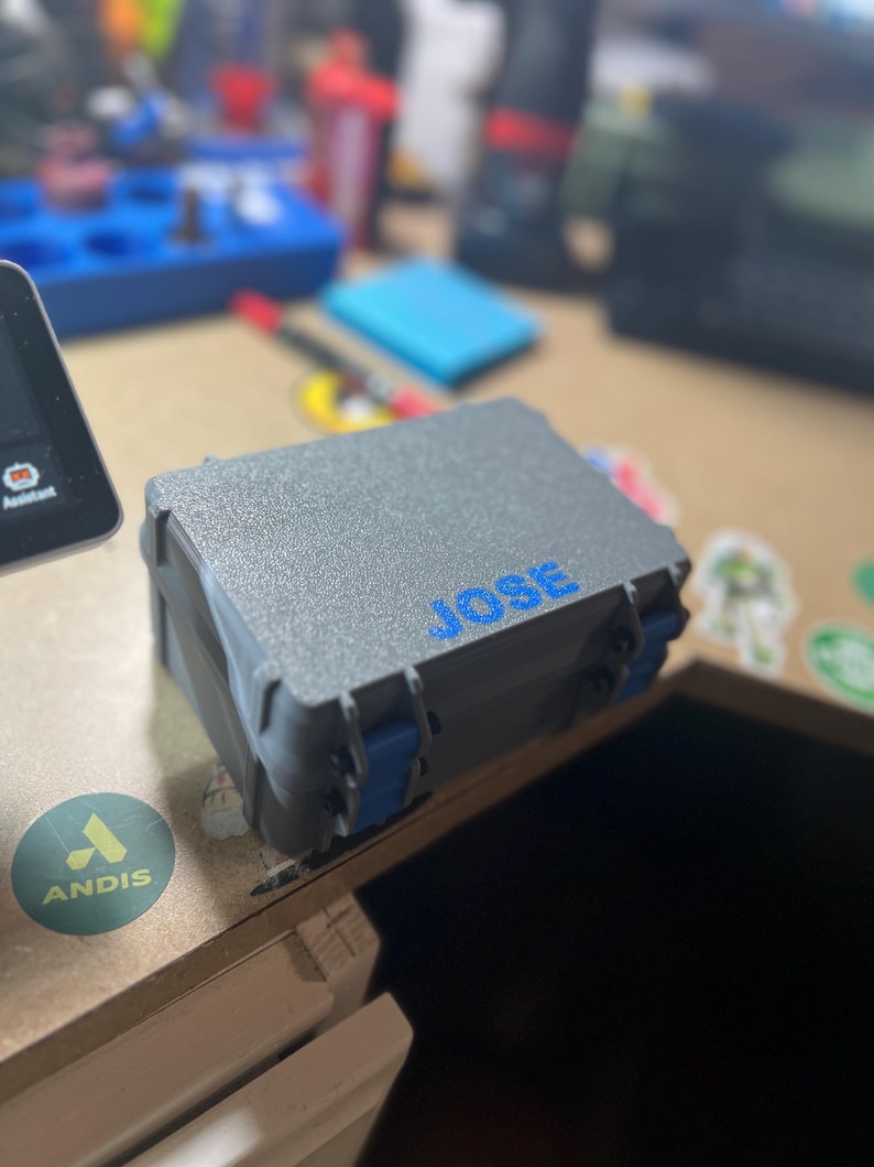 May include: A gray, textured box with blue clasps and the name "JOSE" in blue lettering. The box is closed and appears to be made of durable plastic. The background is blurred, showing other objects on a desk.