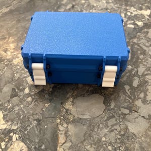 May include: A blue, rectangular, hard-shell case with a textured surface. The case has white latches and black hinges. The case is closed and sits on a mottled grey surface.