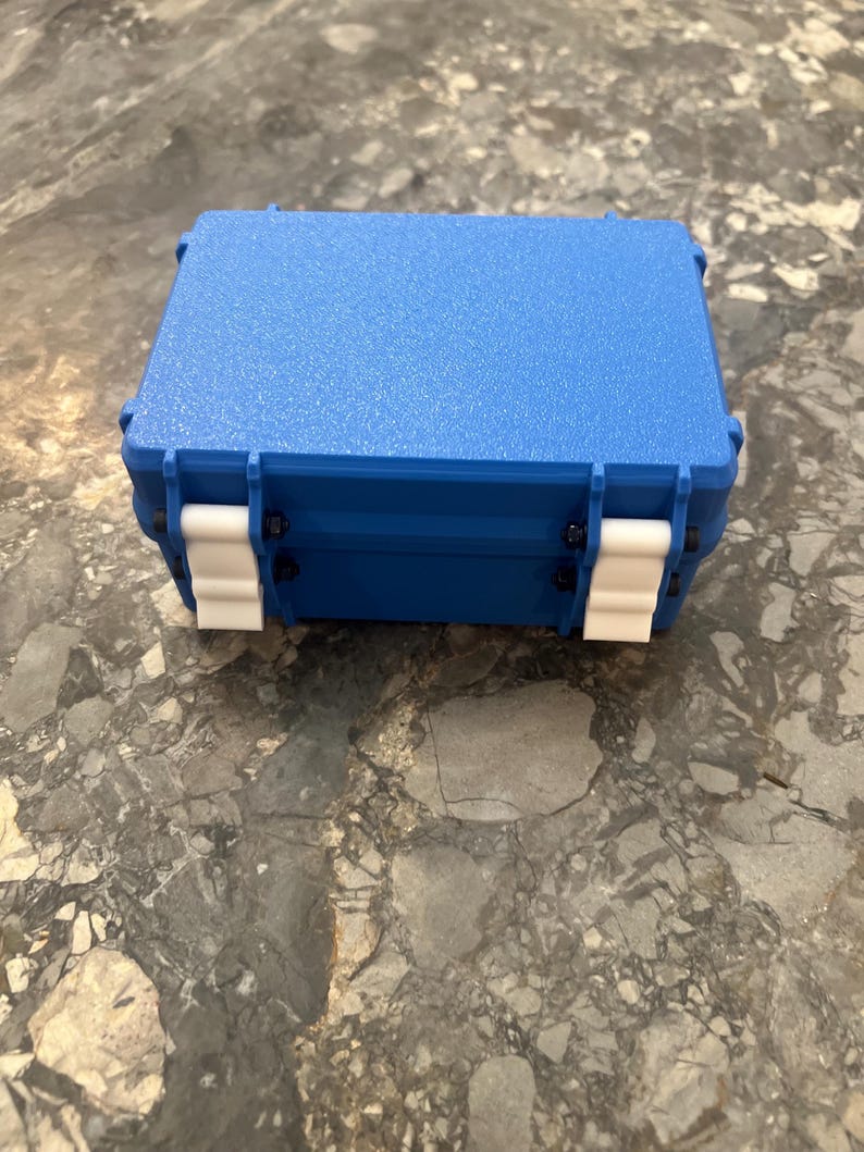 May include: A blue, rectangular, hard-shell case with a textured surface. The case has white latches and black hinges. The case is closed and sits on a mottled grey surface.