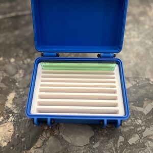 May include: Open blue plastic case with a white interior and multiple slots. The case has a hinged lid and white latches. The interior has a white foam base with multiple slots for storage. A green plastic piece is visible inside the case.