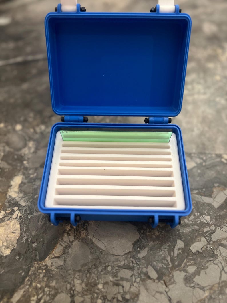 May include: Open blue plastic case with a white interior and multiple slots. The case has a hinged lid and white latches. The interior has a white foam base with multiple slots for storage. A green plastic piece is visible inside the case.