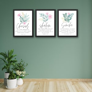 Puede incluir: Tres impresiones artísticas enmarcadas con marcos negros, cada una con diseños florales en acuarela en rosa, dorado y verde. Las impresiones muestran las palabras "Chesed", "Shalom" y "Simcha" con texto bíblico.