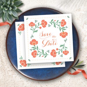 Pode incluir: Dois cartões quadrados "Save the Date" com um design de coroa de flores. Os cartões apresentam flores laranja e folhas verdes, com o texto "Save the Date" em uma fonte cursiva. Os cartões estão em um prato azul escuro.