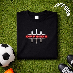 Puede incluir: Camiseta negra con la palabra "OFFSIDE" en un diseño estilizado. La camiseta está sobre una superficie verde con un balón de fútbol y botas de fútbol visibles. La palabra "OFFSIDE" también está escrita en un diseño curvo.