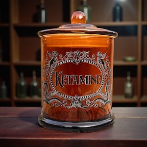 May include: An amber glass apothecary jar with a lid, labelled "KETAMINE" in ornate lettering. The label is framed with decorative scrollwork and cherub figures. The jar is on a wooden surface.