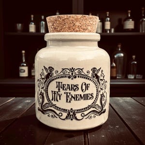 May include: A cream ceramic jar with a cork stopper. The jar has a black ornate design with the text "Tears Of My Enemies" in a decorative oval frame. The background includes a dark wooden surface and a shelf with bottles.