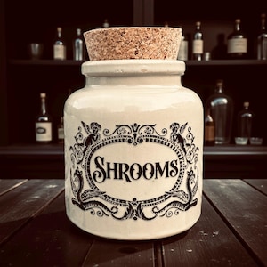 May include: A cream-coloured ceramic jar with a cork stopper. The jar has a black ornate label with the word "SHROOMS" in a decorative font. The label includes angel-like figures and scrollwork. The jar is on a dark wooden surface.