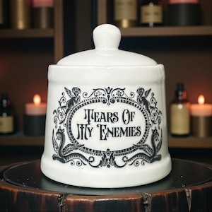 May include: White ceramic jar with a lid, inscribed with "Tears Of My Enemies" in a black, decorative frame. The jar is round and adorned with cherubs and mythical figures. The background features candles and bottles.