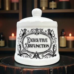 May include: White ceramic jar with lid, featuring the text "EXECUTIVE DISFUNCTION" in a black, ornate oval design. The jar has a round shape and a small knob on the lid. Decorative elements include cherubs and stylized sea creatures.