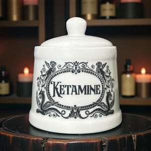 May include: White ceramic apothecary jar with lid, featuring the word "KETAMINE" in a black oval frame with decorative angel and serpent motifs. The jar sits on a dark wooden surface, with candles and bottles in the background.