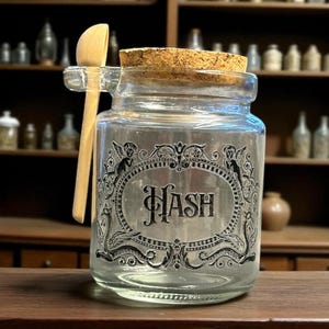 May include: A clear glass jar with a cork lid and a wooden spoon. The jar features a decorative label with the word "HASH" in the centre. The label is surrounded by ornate scrollwork and angel figures. The jar is on a wooden surface.