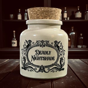 May include: A cream ceramic jar with a cork stopper, labelled "Deadly Nightshade" in a black decorative frame. The jar has ornate black illustrations of cherubs and mythical creatures. The background includes a dark wooden shelf with bottles.