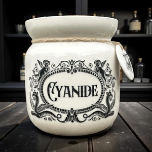 May include: A cream-coloured ceramic jar with a black label reading "CYANIDE". The jar is decorated with cherubs and scrollwork. A rope detail encircles the neck, and a small tag is attached. The jar is on a dark wooden surface.