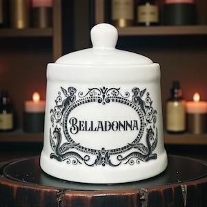 May include: White ceramic apothecary jar with a lid and a decorative oval label that reads "Belladonna" in black script. The label is framed by ornate black illustrations of cherubs and mythical creatures. The jar is displayed on a dark wooden surface.