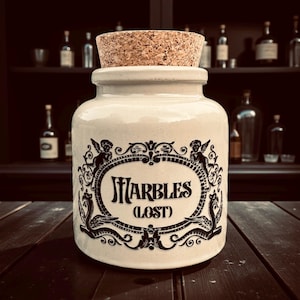 May include: A cream ceramic jar with a cork stopper. The jar features a black ornate design and the words "MARBLES (LOST)". The jar is on a dark wooden surface, with a shelf of bottles in the background.