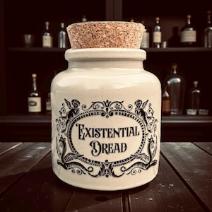 May include: A cream ceramic jar with a cork stopper. The jar features a black ornate design and the words "Existential Dread". The jar is on a dark wooden surface. The background includes shelves with bottles.