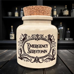 May include: A cream-colored apothecary jar with a cork stopper. The jar features a black ornate label with the words "EMERGENCY SEROTONIN". The label is framed by cherubs and decorative flourishes. The background includes a dark wooden surface and a cabinet with bottles.