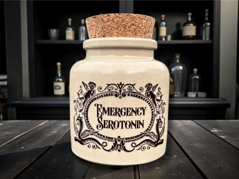 May include: A cream-colored apothecary jar with a cork stopper. The jar features a black ornate label with the words "EMERGENCY SEROTONIN". The label is framed by cherubs and decorative flourishes. The background includes a dark wooden surface and a cabinet with bottles.