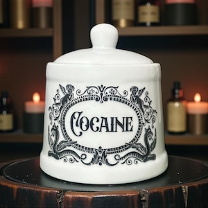 May include: White ceramic apothecary jar with a lid, featuring the word "COCAINE" in a black oval frame with cherubs and scrollwork. The jar is on a dark wooden surface. The background includes candles.
