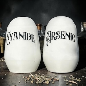 May include: Two white ceramic jars, each with a black gothic label: "Cyanide" and "Arsenic". The jars are oval and rest on a dark wooden surface. Dried herbs are scattered around the base, with glass bottles visible in the background.