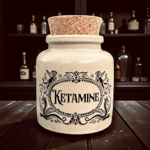 May include: A cream ceramic jar with a cork stopper. The jar has a black ornate design with the word "KETAMINE". The background includes a dark wooden surface and a shelf with bottles.