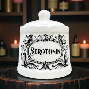 May include: A white ceramic apothecary jar with a lid, featuring the word "Serotonin" in a decorative black oval. The jar is adorned with black illustrations of cherubs and mythical creatures. The background includes candles and apothecary bottles.