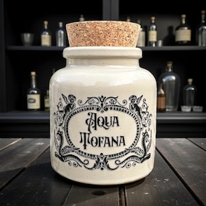 May include: Cream ceramic jar with a cork stopper, displaying black ornate lettering and illustrations. The text reads "Aqua Tofana" within a decorative oval frame. The jar is on a dark wooden surface.