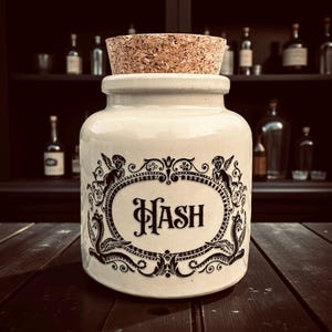 May include: Cream ceramic jar with a cork stopper, displaying the word "HASH" within an ornate oval frame. The frame is decorated with cherubic figures and scrollwork. The jar rests on a dark wooden surface.
