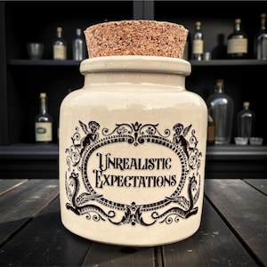 May include: A cream-colored ceramic jar with a cork stopper. The jar features a black ornate design with the words "Unrealistic Expectations" in a decorative oval. The background includes a dark wooden surface and a shelf with bottles.