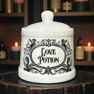 May include: A white ceramic jar with a lid, displaying the words "Love Potion" within a decorative oval frame. The frame is embellished with cherubs and ornate scrollwork in black. The jar is set against a backdrop of candles and apothecary bottles.