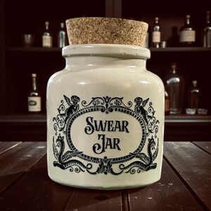 May include: Cream ceramic jar with a cork lid. The jar has a black ornate design with the words "SWEAR JAR". The design includes cherubs and decorative flourishes. The jar is on a dark wooden surface. The background shows bottles.