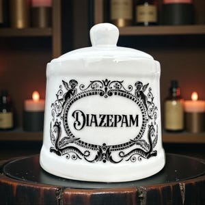 May include: A white ceramic apothecary jar with a lid, featuring the word "DIAZEPAM" in a decorative oval frame. The frame is adorned with cherubs and ornate scrollwork in black. The jar is displayed on a dark surface.