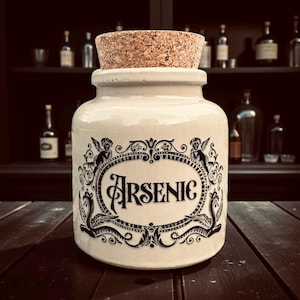 May include: An off-white ceramic jar with a cork stopper, labelled "Arsenic" in black, decorative lettering. The jar is decorated with black scrollwork and cherub motifs. It sits on a dark wooden surface, with bottles in the background.