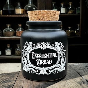 May include: A black ceramic jar with a cork stopper. The jar features white ornate lettering that reads "Existential Dread". The design includes cherubs and decorative flourishes. The jar is displayed on a wooden surface.