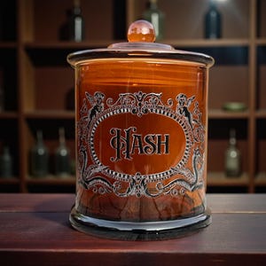 May include: Amber glass apothecary jar with a lid and the word "HASH" in an oval frame. The jar has a decorative design with cherubs and scrollwork. The jar is on a wooden surface.