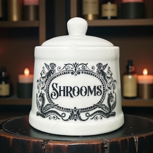 May include: White ceramic jar with a lid, displaying the word "SHROOMS" in a black oval frame, decorated with angel and serpent illustrations. The jar sits on a dark wooden surface, with candles and bottles in the background.
