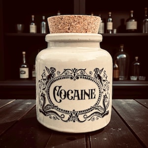 May include: A cream-coloured ceramic jar with a cork stopper. The jar features a black decorative border and the word "COCAINE" in a stylized font. The background shows a dark wooden shelf with various bottles.