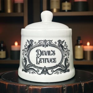 May include: White ceramic jar with a lid, displaying the text "Devil's Lettuce" within a decorative oval frame. The frame is embellished with angel and swan illustrations in black. The jar is placed on a dark surface.
