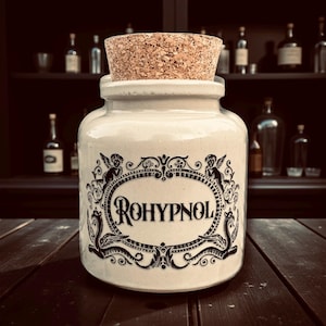 May include: A cream ceramic apothecary jar with a cork stopper. The jar has a black decorative label with the word "ROHYPNOL" in a vintage font. The label is framed by ornate scrollwork and cherub figures. The background includes a dark wooden shelf with bottles.