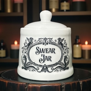 May include: A white ceramic "Swear Jar" with a lid. The jar has a black ornate design with the words "Swear Jar" in the centre. The design includes cherubs and decorative elements. The jar is on a dark surface.