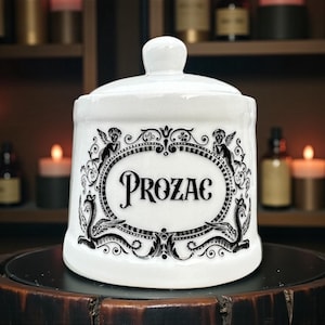 May include: White ceramic apothecary jar with a lid, featuring the word "PROZAC" in a black oval frame. The frame is decorated with cherubs and ornate scrollwork. The jar sits on a dark wooden surface.