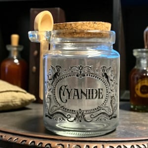 May include: A clear glass jar with a cork stopper and a wooden spoon. The jar has a label with the word "CYANIDE" in a decorative oval frame. Other apothecary bottles are in the background.